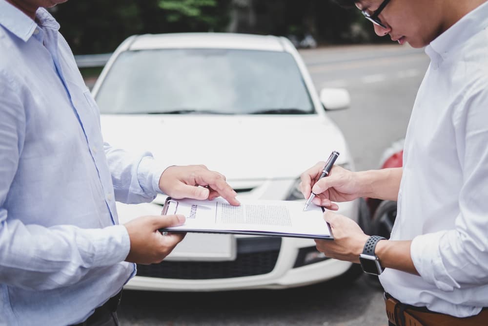 An insurance agent inspects a damaged car while a customer signs a report claim form after a traffic accident, representing the insurance process.