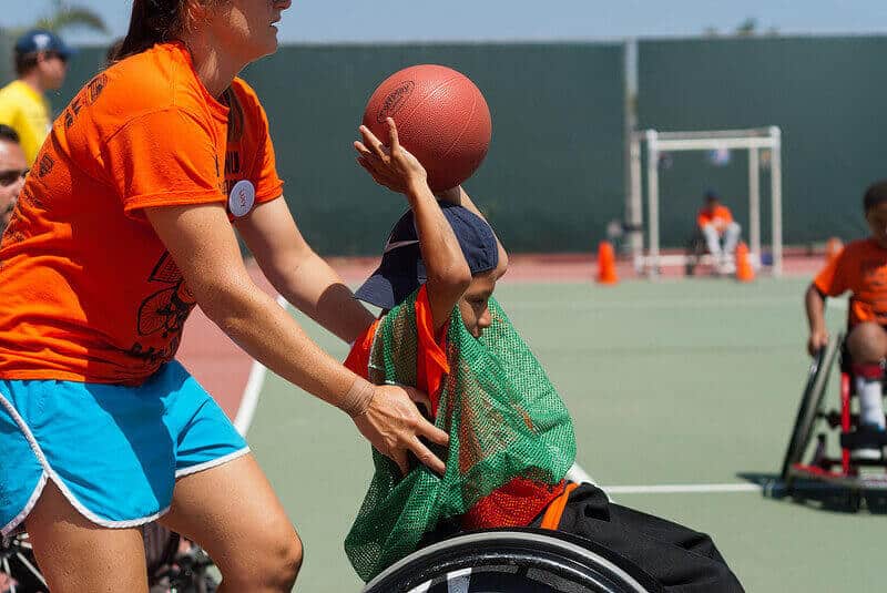child with a spinal cord injury playing basketball in a wheelchair with help of caretaker