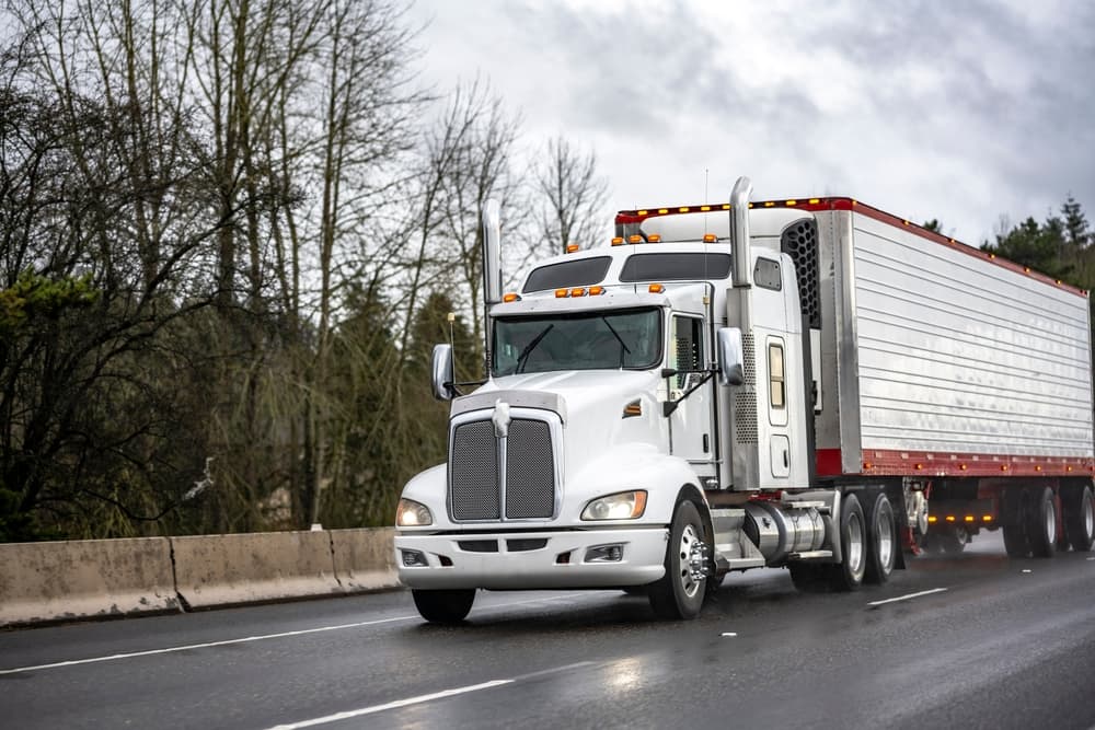 Gray long-haul big rig with extended cab and reefer trailer, transporting cargo on a rain-dusted twilight highway road.