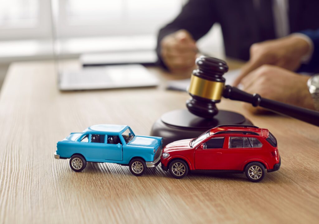 In a courtroom setting, miniature crashed cars rest on a table, accompanied by a gavel and two toy car models on the desk. Symbolizing legal services, civil court trials, vehicle accident case studies, and insurance coverage concepts.