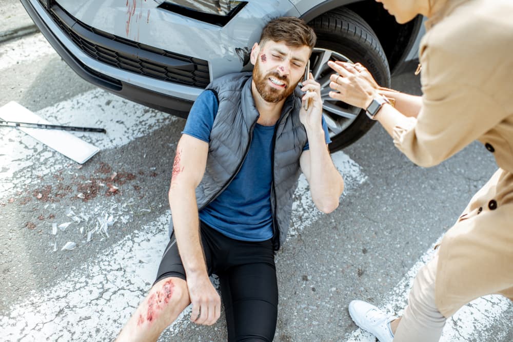Injured man and woman driver discussing the situation on the phone near a damaged car at a pedestrian crossing after the accident.