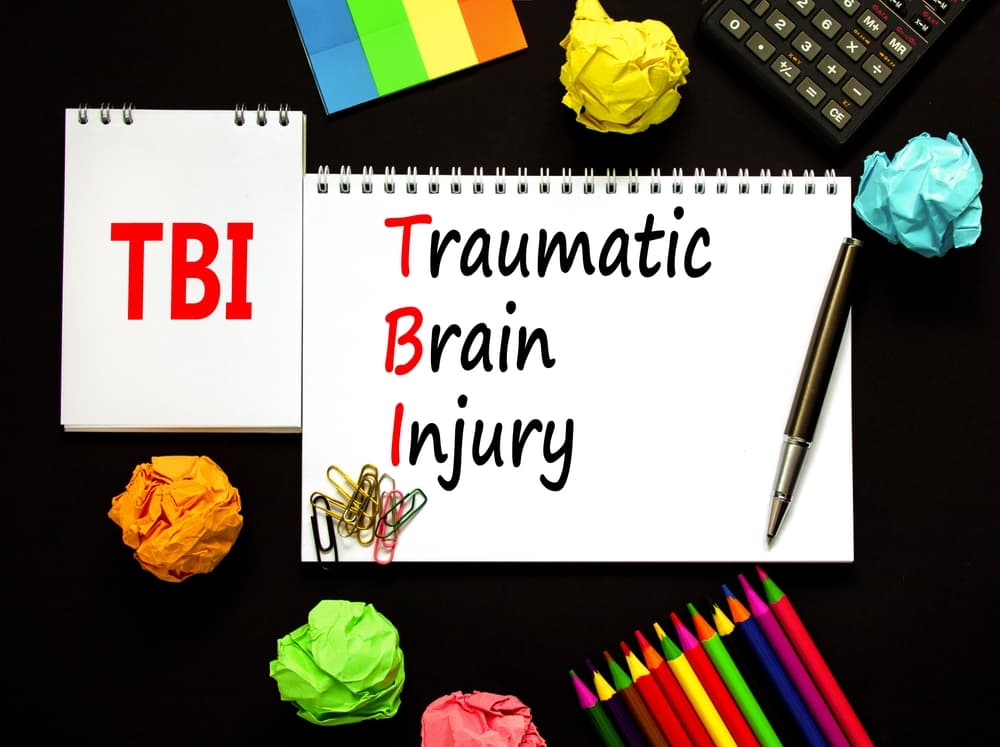 Symbolic representation of TBI (Traumatic Brain Injury) with words on a white note against a black background. Medical concept with pencil and pen, offering copy space.