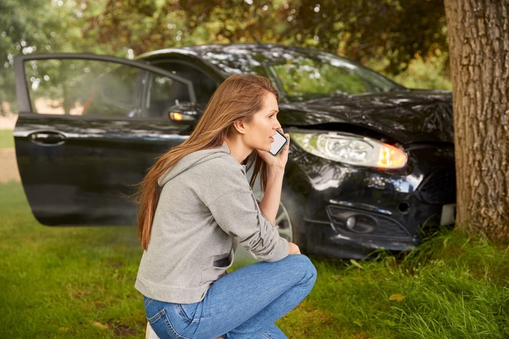 Woman examining car crash near a tree, assessing damage, and calling for emergency assistance.