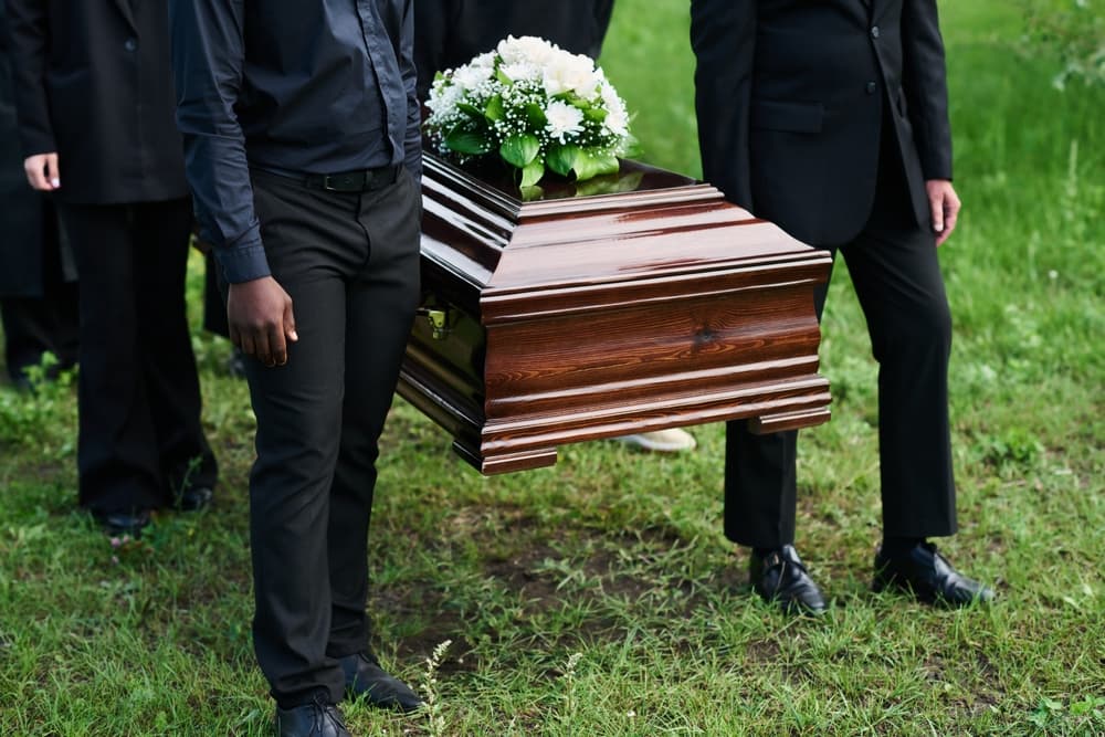 A diverse group of men, dressed in black, carrying a wooden coffin adorned with white flowers on the closed lid.