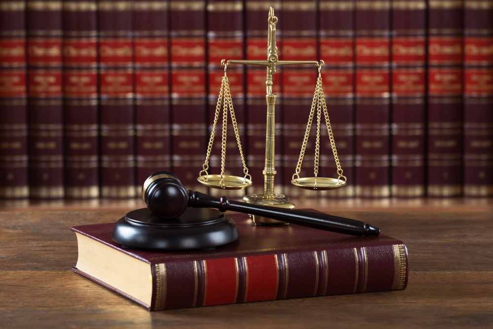 The image features a legal-themed still life composition. In the foreground, a wooden gavel rests on a thick, leather-bound book with a red and gold cover. Next to the gavel, there is a set of golden scales of justice. The background is filled with rows of law books, identifiable by their uniform size, leather bindings, and gold lettering on the spines, reinforcing the legal context of the scene. The overall setting conveys a sense of authority, law, and order.