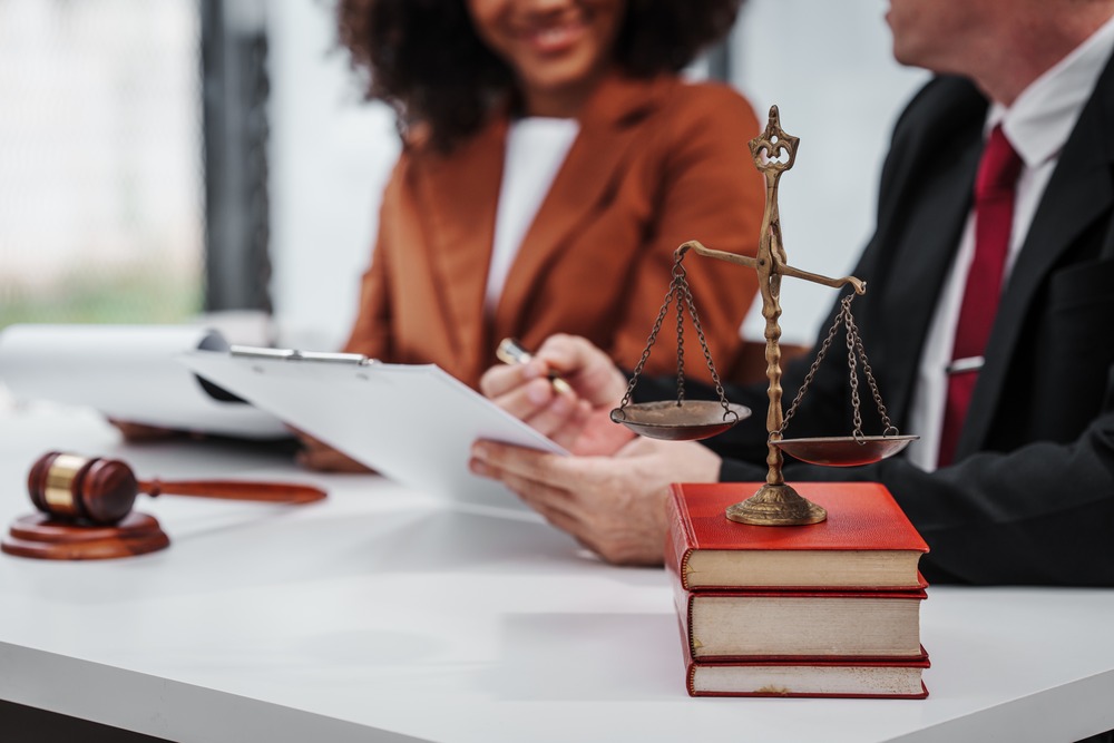 Two professionals are seated at a table, engaged in a discussion. One is holding a clipboard and reviewing documents, while the other listens attentively. On the table, there are a gavel, a stack of red books, and a decorative brass scale of justice, symbolizing law and justice. The atmosphere suggests a legal consultation or a business meeting. The background is softly blurred, focusing attention on the interaction and the items on the table.